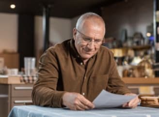 Een bejaarde zit aan een tafel en kijkt naar een stuk papier dat voor hem ligt.