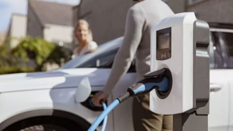 Een close-up van een oplaadpunt, een man die een blauwe slang vasthoudt om zijn witte auto op te laden en een dame die in de auto stapt vanaf de passagiersstoel. 