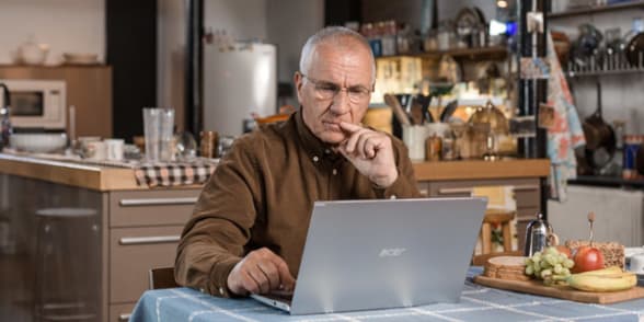 Een oudere man zit aan een tafel, gebruikt een laptop en concentreert zich op zijn werk.