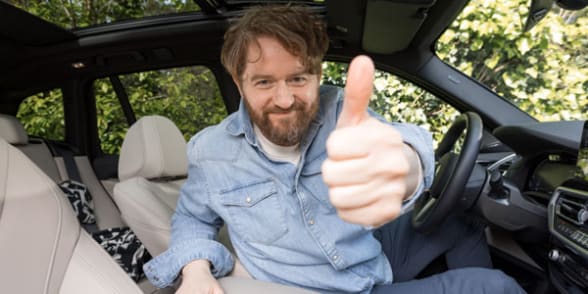 Een man met een baard glimlacht en steekt zijn duim omhoog vanuit zijn auto.