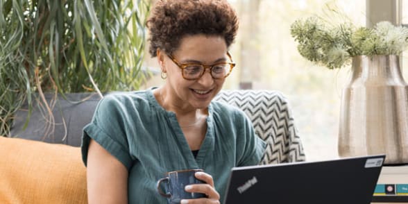 Een vrouw met een bril glimlacht terwijl ze een kop koffie vasthoudt, zittend op een bank met een laptop.