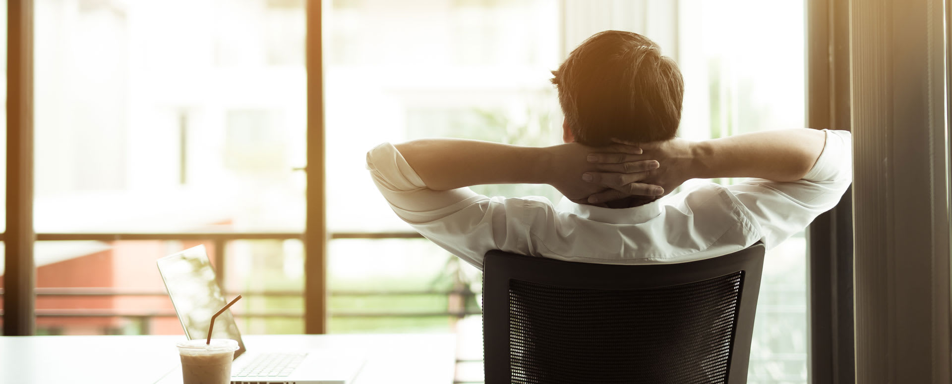 Un homme est assis sur une chaise, les mains derrière la tête, dans son bureau, et regarde la vue par la fenêtre.