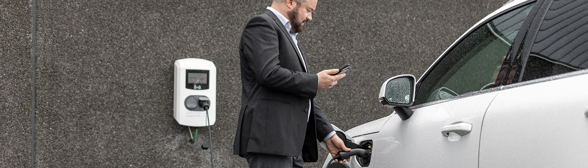 Un homme tient et regarde son téléphone portable. De l&#039;autre main, il tient la pompe électrique pour recharger sa voiture.